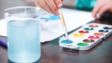 يد تحتوي على فرشاة رسم مغمورة في لون أزرق من علبة ألوان مائية بجانب كوب من الماء. تظهر ورقة بيضاء تحمل رسومات ملونة في الخلفية.