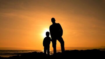 رجل وطفل يقفان silhouetted أمام غروب الشمس الجميل على الساحل، مع ألوان دافئة في السماء تعكس لحظة عاطفية.