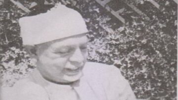 صورة تاريخية لرجل مسن يرتدي قبعة بيضاء وقميص فاتح، يظهر في وضعية تفكير. الخلفية تتضمن نباتات خضراء، مما يضفي شعوراً بالطبيعة والهدوء. تعكس الصورة أجواءً من الزمن الماضي، مما يجعلها جذابة للمهتمين بالتاريخ والثقافة.