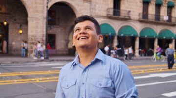 رجل شاب يبتسم نظرًا إلى السماء في شارع مزدحم مع مباني تاريخية في الخلفية. يظهر الناس في الخلفية، بما في ذلك بعض الدراجات.