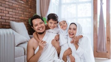 عائلة سعيدة تتكون من أب وأم وطفلين، يرتدون ملابس بيضاء، يتواجدون في غرفة مع جدران من الطوب. الأب يحتضن أطفاله، بينما الأم تبتسم. الجو مريح ومشمس.