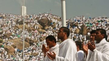 مجموعات كبيرة من الحجاج يرتدون ملابس الإحرام البيضاء، يقفون في موقع مزدحم أثناء أداء شعائر الحج، مع تلال وصخور في الخلفية وجو روحاني.