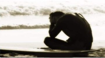 شخص جالس على الشاطئ، يرتدي بدلة غوص، يظهر في وضع تفكير عميق مع الأمواج في الخلفية، يعكس شعور التأمل والهدوء.