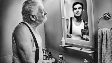 رجل مسن يقف أمام مرآة في حمام، يعكس صورته الشابّة في المرآة. يظهر الرجل بملابس داخلية بيضاء، مع أشرطة على الكتفين، ويبدو عليه التأمل والحنين. الحمام يحتوي على منتجات عناية شخصية ومناشف. الصورة بالأبيض والأسود تعكس مشاعر الزمن والتغيير.