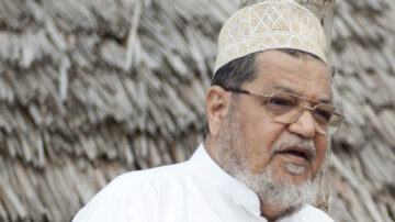 رجل مسن يرتدي عمامة تقليدية ونظارات، يتحدث في مجال مفتوح مع خلفية من السقف المصنوع من القش.