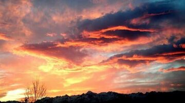 غروب الشمس فوق الجبال المغطاة بالثلوج، سماء ملونة بألوان دافئة مثل البرتقالي والأحمر، سحب داكنة تتداخل مع الأضواء الساطعة، مشهد طبيعي مذهل يعكس جمال الطبيعة.