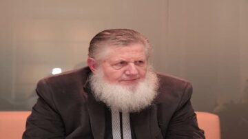 رجل مسن ذو لحية بيضاء، يرتدي بدلة داكنة، يجلس في غرفة ذات جدران زجاجية. يبدو عليه التركيز والتفكير.