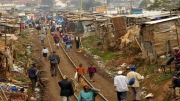 مشهد لشارع مزدحم في حي فقير، يظهر فيه الناس يتجولون بين أكواخ مصنوعة من الصفيح، مع وجود عربات وأكوام من المواد. يُظهر المشهد الحياة اليومية والتحديات الاقتصادية في المناطق الحضرية.