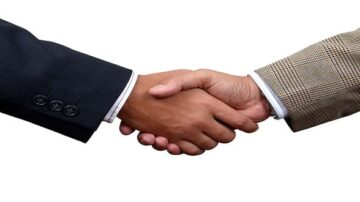 مصافحة بين شخصين يرتديان بدلتين، واحدة داكنة والأخرى مزخرفة. تعكس الصورة روح التعاون والاتفاق.