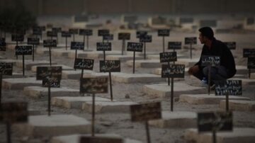 رجل يجلس في مقبرة محاطة بشواهد قبور تحمل أسماء وتواريخ، يعكس لحظة من التأمل والحزن وسط الأجواء القاتمة.