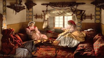 نساء جالسات في غرفة تقليدية مزينة بألوان دافئة. تظهر الأثاث المزخرف والسجاد الفاخر، مع نافذة تعكس الضوء الطبيعي. تعكس الصورة الثقافة والتراث العربي.