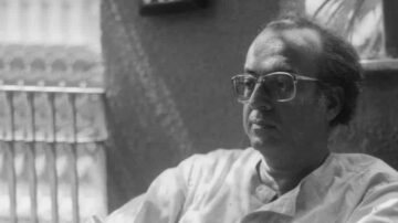 رجل يرتدي نظارات يجلس في غرفة مضاءة بشكل جيد، مع تعبير جاد على وجهه. خلفية الصورة تظهر تفاصيل معمارية بسيطة، مما يضيف عمقًا إلى المشهد.