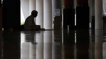 رجل يجلس في وضع الصلاة داخل مسجد، بظلال مميزة تعكس الضوء على الأرض اللامعة، مما يعكس أجواء السكون والروحانية.