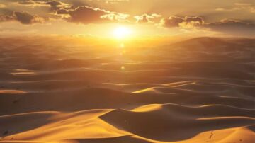 غروب الشمس فوق الكثبان الرملية، مع سماء مليئة بالسحب. الضوء الذهبي يضيء المناظر الطبيعية الصحراوية، مما يخلق مشهداً ساحراً من الألوان الدافئة.