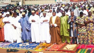 مجموعة كبيرة من الرجال يرتدون ملابس تقليدية مختلفة يؤدون الصلاة في تجمع جماهيري، مع وجود سجاد صلاة ملون تحت أقدامهم.