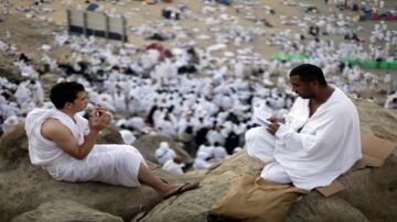 رجلان يرتديان ملابس الإحرام يجلسان على صخرة في مكان مزدحم بالحجاج. الخلفية مليئة بالحجاج الذين يرتدون الملابس البيضاء، مما يدل على أجواء الحج.