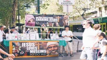 منصة دعوية في منطقة مزدحمة، تحتوي على لافتات تحمل رسائل عن المسيحية، ووجود أشخاص يتجولون حولها. تبرز اللافتات عبارة "يسوع يحب المسلمين". تظهر مركبة ترام في الخلفية وأشخاص مختلفون يمشون في اتجاهات متعددة.