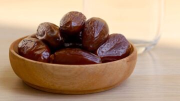 تمر طازج في وعاء خشبي صغير، مع زجاجة ماء خلفه. تعكس الحبات لونها البني اللامع وتظهر قوامها الناعم.