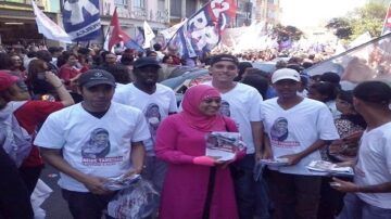 مجموعة من الأشخاص في تظاهرة حاشدة، يرتدون قمصانًا بيضاء تحمل صورة امرأة. يحملون منشورات ويبتسمون، بينما تتواجد خلفهم لافتات وأعلام. تظاهر الحشود في الشوارع تعكس روح النشاط والمشاركة.