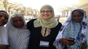 امرأة ترتدي حجابًا وتبتسم وسط ثلاث نساء يرتدين ملابس تقليدية، في موقع ديني. تظهر معالم معمارية تقليدية في الخلفية، مما يشير إلى مكان مقدس.