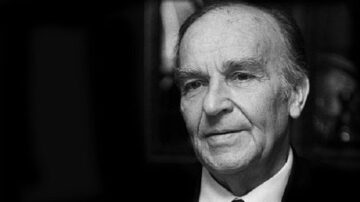 صورة لرجل مسن ذو شعر رمادي، يرتدي بدلة داكنة، يبدو عميق التفكير. خلفية الصورة غير واضحة وتضفي طابعًا دراميًا.