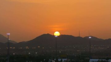 غروب الشمس فوق الجبال، مع توهج برتقالي وأصفر في السماء، وأضواء كاشفة تضيء المنطقة السكنية في الأسفل.