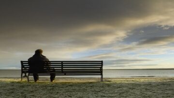 رجل يجلس على مقعد في حديقة مطلة على بحر هادئ تحت سماء غائمة، مع انعكاسات ضوء الشمس على الماء.