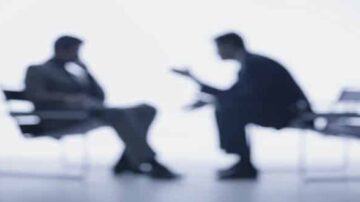 محادثة بين رجلين يجلسان على كراسي، حيث يظهران بشكل ضبابي في الخلفية. تعبيرات الوجه غير واضحة، مما يعكس تفاعلًا عميقًا.