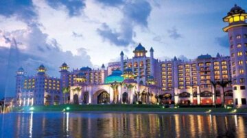 فندق فاخر بتصميم معماري مميز، يضيء بألوان دافئة تحت السماء الزرقاء الداكنة. المبنى يحتوي على أبراج مزينة وأضواء تعكس جماله على سطح الماء. يحيط به مناظر طبيعية خضراء.