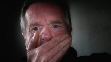 رجل مسن يغطي فمه بيده، مع تعبير وجه يعكس الخوف أو الصدمة، خلفية مظلمة تعزز الشعور بالقلق.