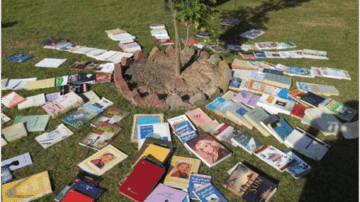 كتب متنوعة مرتبة حول شجرة صغيرة في حديقة، مع تنوع في الألوان والتصاميم. الكتب تتضمن روايات وكتب تعليمية، مما يخلق مشهداً جذاباً للطبيعة والثقافة.