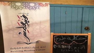 شعار نادي الكتب "إحياء ثقافة القراءة" أمام لوحة طباشير تعرض تفاصيل حدث حكمة مطلع، بتاريخ 5 فبراير 2015، في مكان ذو أبواب زرقاء.