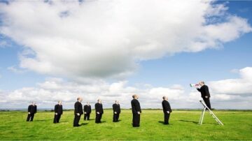 مجموعة من الرجال يرتدون بدلات سوداء يقفون في صفوف على أرض خضراء، بينما يقف رجل آخر على سلم ويستخدم مكبر صوت. السماء مليئة بالغيوم البيضاء. الصورة تعبر عن القيادة والتوجيه.