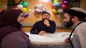 شخصان يجلسان على طاولة مزينة بالبالونات الملونة، حيث تتحدث امرأة محجبة مع رجل بينما ينظر إليهما رجل آخر يجلس بينهما. تعكس الصورة لحظة من الفرح والاحتفال.