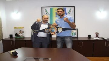 رجلان يقفان في مكتب يحمل أحدهما حزام بطولة، معروضًا بفخر. الخلفية تحتوي على صور وأوراق. الجو احتفالي يعكس إنجازات رياضية.