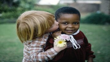 طفلان يلعبان في حديقة، طفل ذو شعر أشقر يقبل طفلة ذات بشرة داكنة على خدها. الطفلة تحمل زهورًا ملونة، كلاهما مبتسم ويعبران عن البراءة والصداقه.