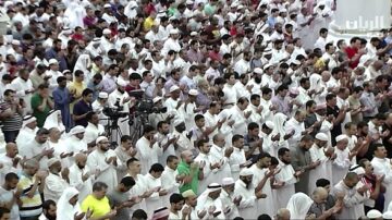 مئات من المصلين يؤدون الصلاة في الحرم المكي، يرتدون ملابس بيضاء، مع التركيز على الأجواء الروحانية والعبادة.