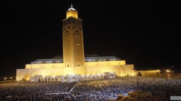 مشهد لجامع الحسن الثاني في الدار البيضاء، المغرب، أثناء صلاة جماعية. يظهر المسجد الكبير مضاءً بأضواء دافئة، مع وجود حشود كبيرة من المصلين في الساحة المحيطة. يبرز المنارة العالية للمسجد في الخلفية، مما يضيف إلى جمال المعلم المعماري.