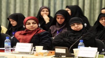 مجموعة من النساء المحجبات يجلسن في قاعة مؤتمر، بعضهن يرتدين سماعات الأذن. تظهر تعبيرات مختلفة على وجوههن، مما يعكس تفاعلهن مع المحتوى المقدم. في الخلفية، توجد ستائر خضراء، ومائدة مزينة بأكواب ومياه.