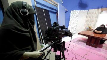 امرأة ترتدي حجابًا وتجلس أمام طاولة أثناء قراءة نص، بينما امرأة أخرى ترتدي سماعات وتحمل كاميرا فيديو لتصوير المشهد. الخلفية تحتوي على ستائر وألوان مريحة.