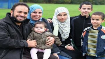 عائلة مكونة من ستة أفراد تتواجد في حديقة. الأب مبتسم ويحتضن ابنته الرضيعة، بينما تجلس الأم بجانبه. الأطفال الأربعة، يتراوح أعمارهم بين الرضيع والمراهق، يظهرون بملابس متنوعة. الخلفية تظهر مساحة خضراء مع شجرة. الصورة تعكس لحظة سعيدة للعائلة.