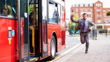 رجل يرتدي بدلة يجري نحو حافلة حمراء تتوقف عند الرصيف، بينما يظهر الركاب في الداخل. تعكس الصورة مشهد الحياة اليومية في المدينة.