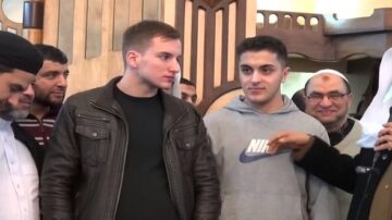 مجموعة من الرجال في مسجد، يتحدث أحدهم في ميكروفون بينما ينظر الآخرون إليه. شخص يرتدي سترة جلدية، وآخر في سترة رياضية. تعبيرات الوجوه تشير إلى التفاعل والمودة.