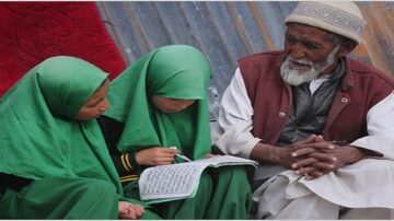 امرأة مسنّة مع لحية يجلس بجوار فتاتين ترتديان حجابًا أخضر، يكتبن في دفتر. الخلفية تحتوي على جدار معدني ومجموعة من الأقمشة الحمراء. تعكس الصورة التفاعل بين الأجيال والتعلم.