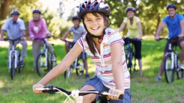 فتاة صغيرة ترتدي خوذة وتبتسم أثناء ركوب دراجة في حديقة، مع مجموعة من الأشخاص البالغين خلفها على دراجات. تعكس الصورة أجواء ممتعة ونشطة في الهواء الطلق.