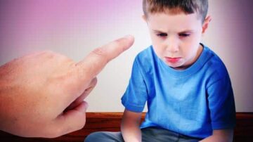 صورة لصبي صغير يجلس على الأرض ويبدو حزينًا، بينما توجد يد تشير إليه من فوق، مما يعكس شعورًا باللوم أو التأنيب.