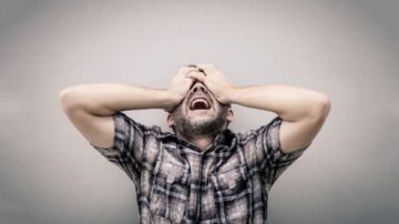 رجل يضع يديه على وجهه ويبدو في حالة من الإحباط أو الضيق. يرتدي قميصًا مزخرفًا ويظهر الخلفية بلون رمادي. تعبر تعابيره عن الشعور بالضغط أو الاستسلام.