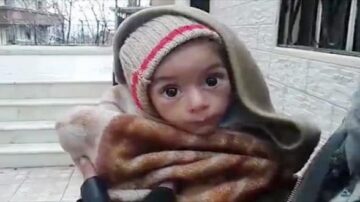 طفل صغير wrapped in a blanket, wearing a knitted hat, with large expressive eyes, standing outside near a building.
