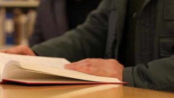 رجل يقرأ كتابًا مفتوحًا على طاولة، تظهر يداه وهو يتصفح الصفحات، مع خلفية مكتبة ضبابية.