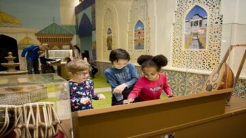 أطفال يستكشفون أنشطة تفاعلية في معرض ثقافي، مع خلفية مزينة بألوان زاهية وأشكال معمارية تقليدية. يُظهر الصورة ثلاثة أطفال يتفاعلون مع أدوات موسيقية، مما يعكس روح التعلم والتفاعل في بيئة تعليمية ممتعة.